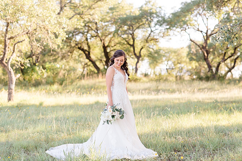 bride holds bouquet to side and looks down at hem of wedding dress in field