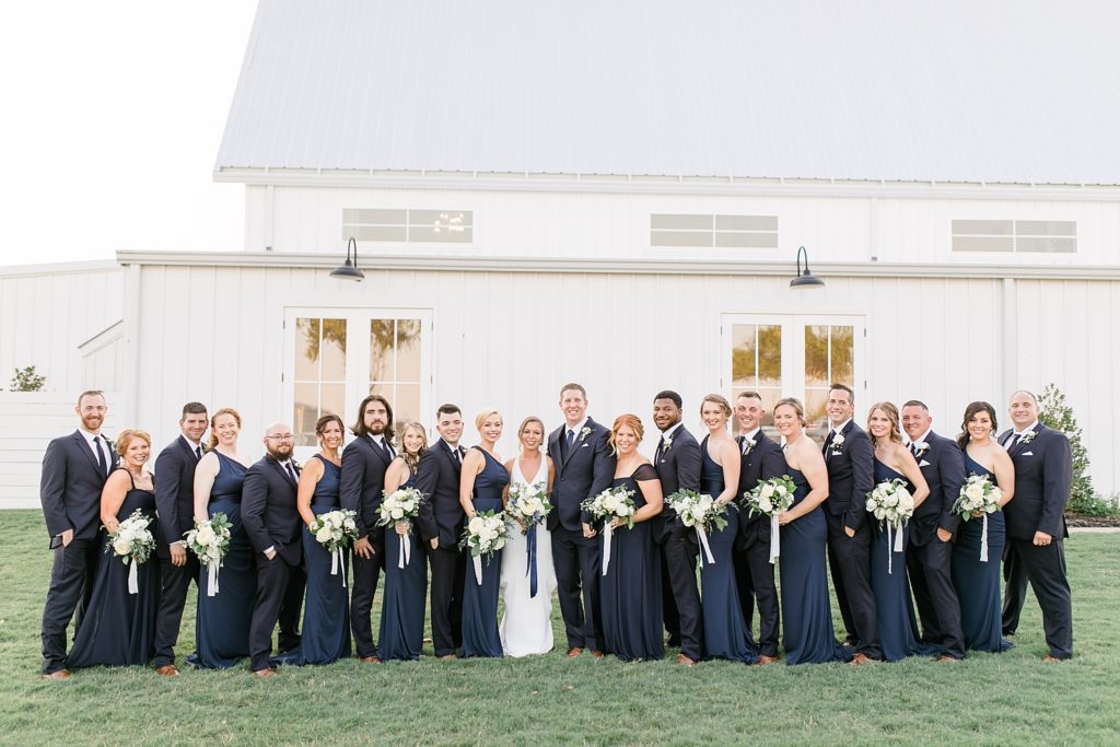 bridal party in navy gowns and black suits pose outside The Nest at Ruth Farms