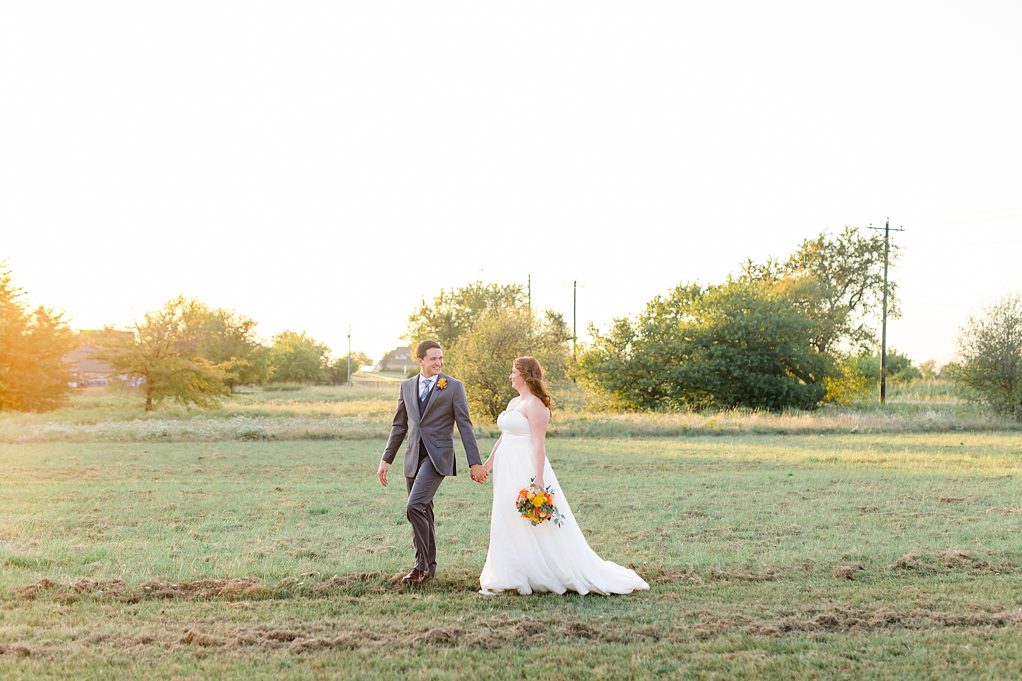 bride and groom walk through field at sunset
