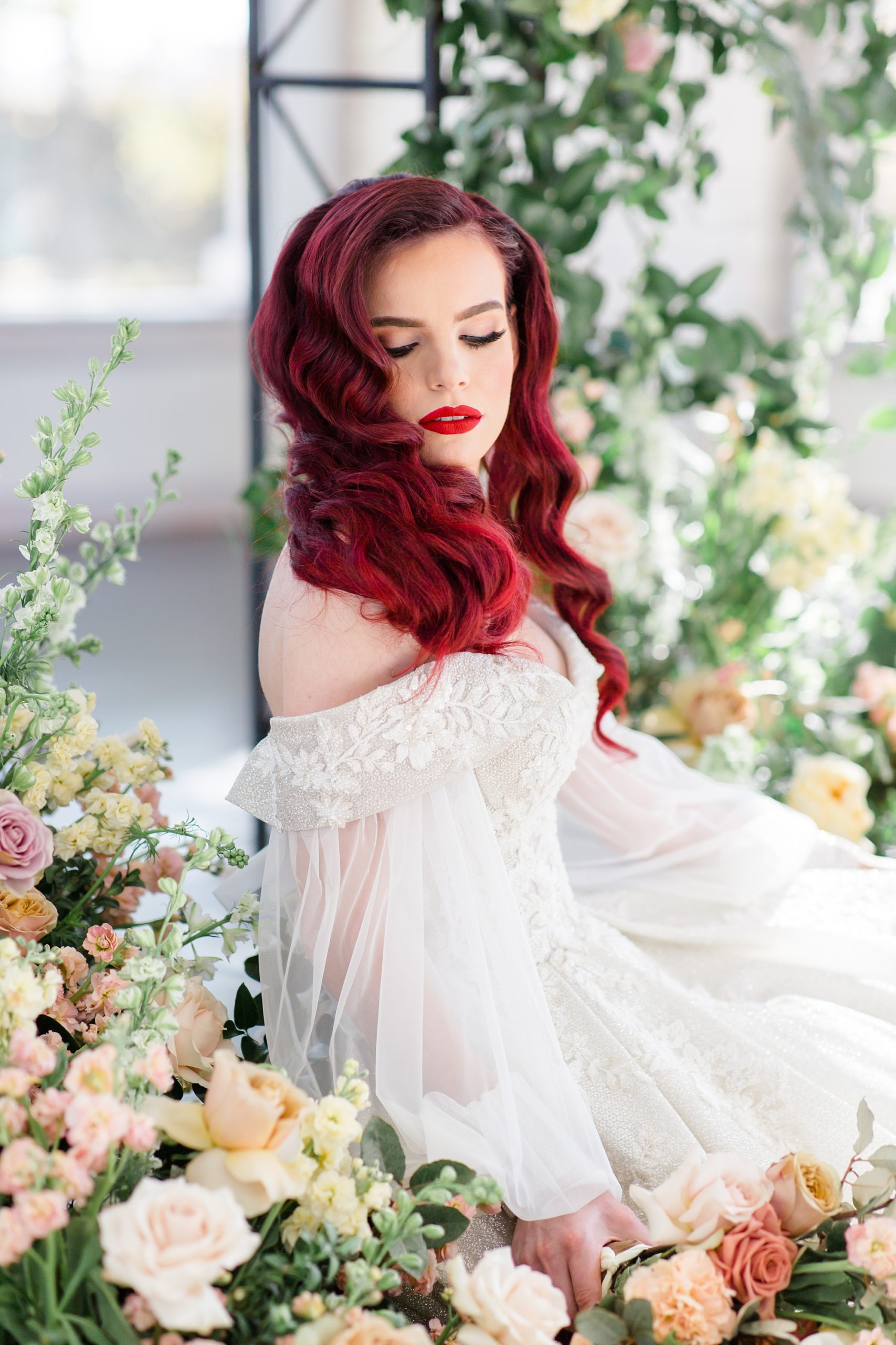 bride with red hair looks over shoulder while sitting amongst flowers by Gothic arch