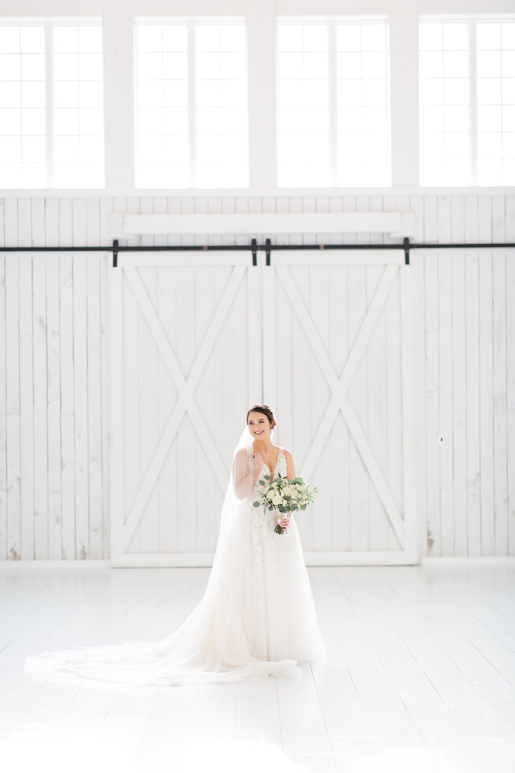 bride-to-be holds bouquet of all white flowers during portraits by barn doors in the White Sparrow