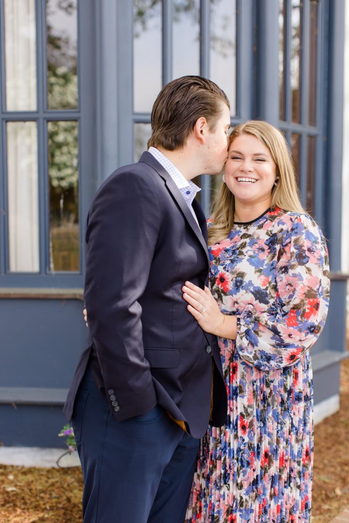 groom kisses fiancees cheek by blue window at Dallas Arboretum