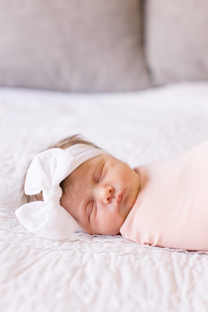 baby in white bow lays on parents' bed during Dallas lifestyle newborn portraits