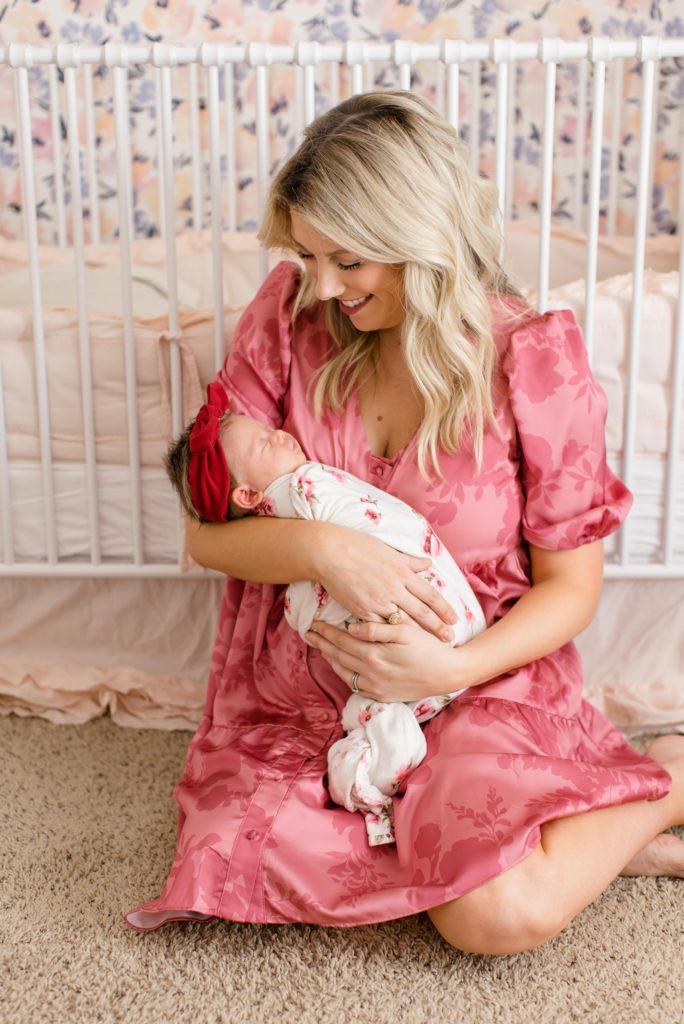 mom holds baby girl in front of crib during Dallas lifestyle newborn portraits