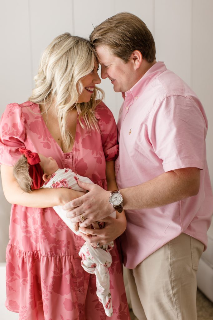 parents stand touching foreheads while holding baby girl 