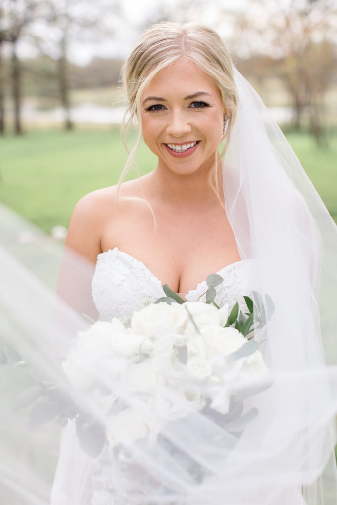 bride smiles holding all-white bouquet with veil draped in front of her