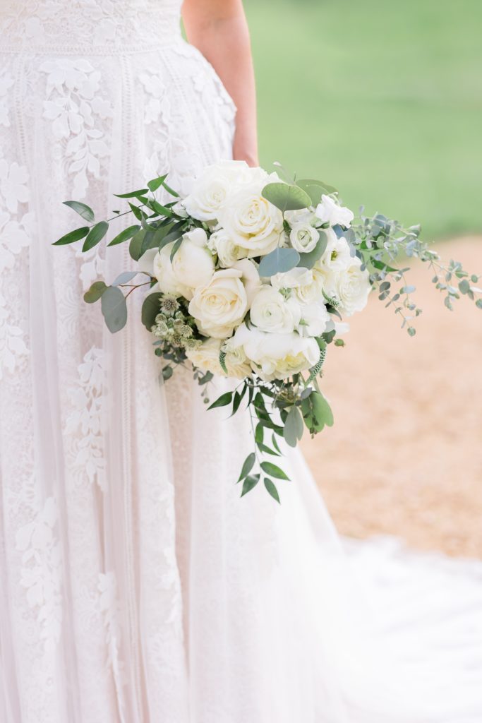 bride holds bouquet of white roses and green leaves at her side 