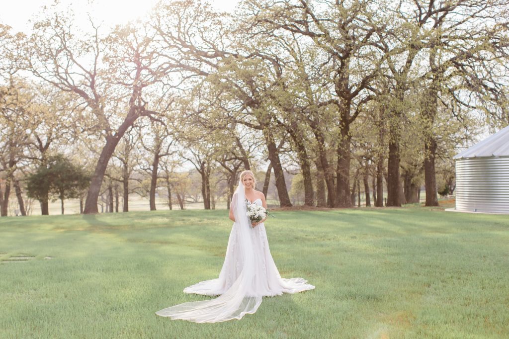 bride stands in meadow with veil wrapped around her shoulders