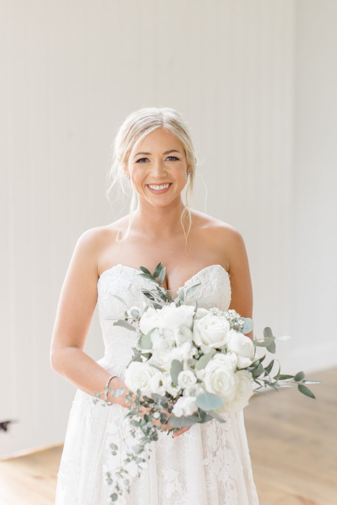 bride holds bouquet of white flowers in front of white wall at Oak + Ivy