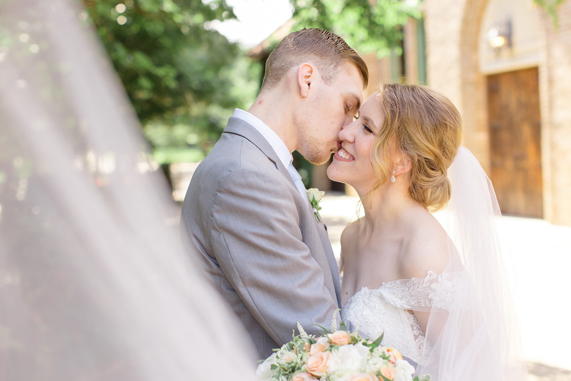 groom kisses bride's cheek while veil lifts around them
