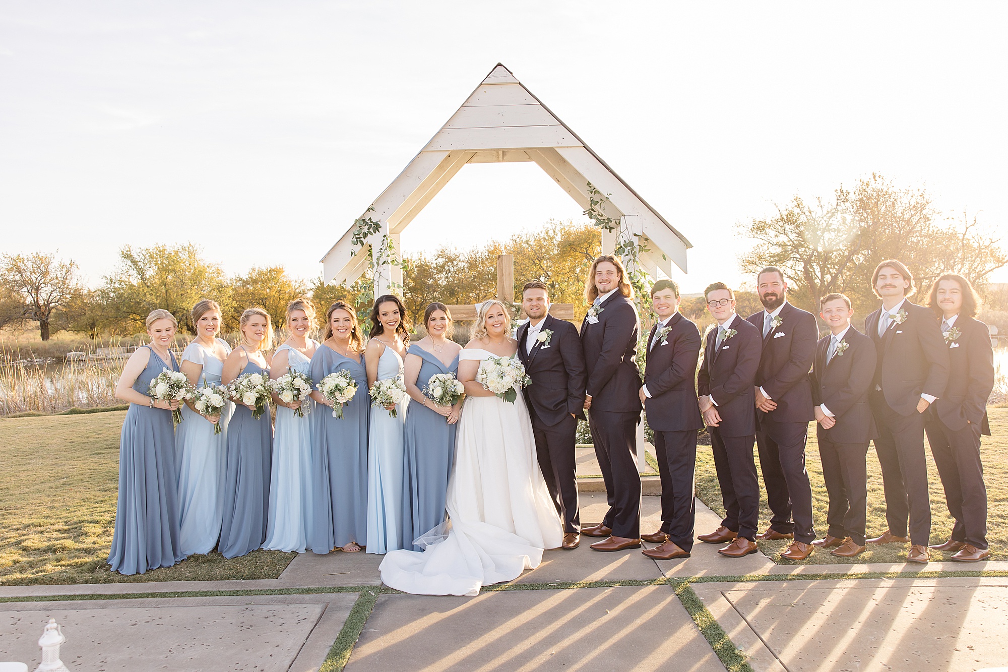 bride and groom stand with wedding party at wooden arbor 