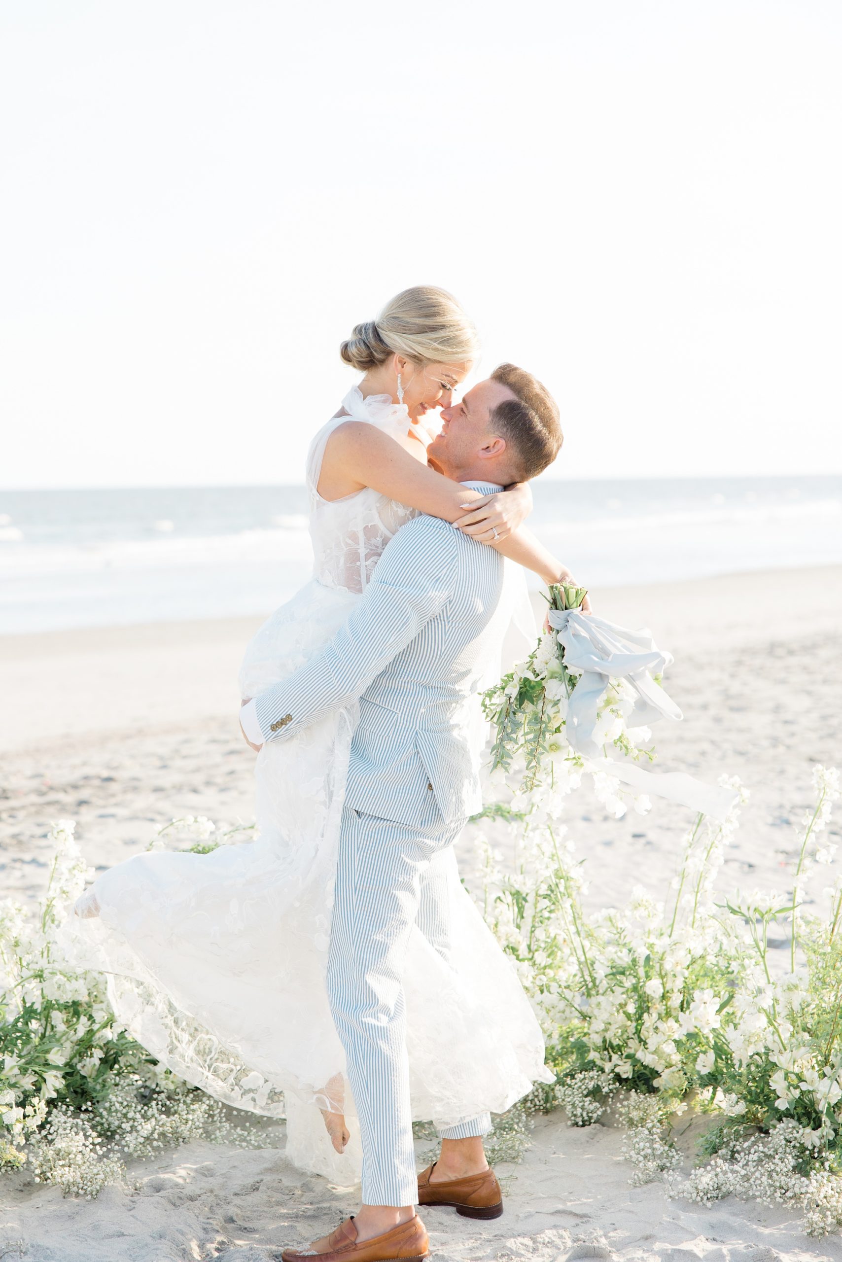 groom in light blue jacket lifts bride up standing on beach