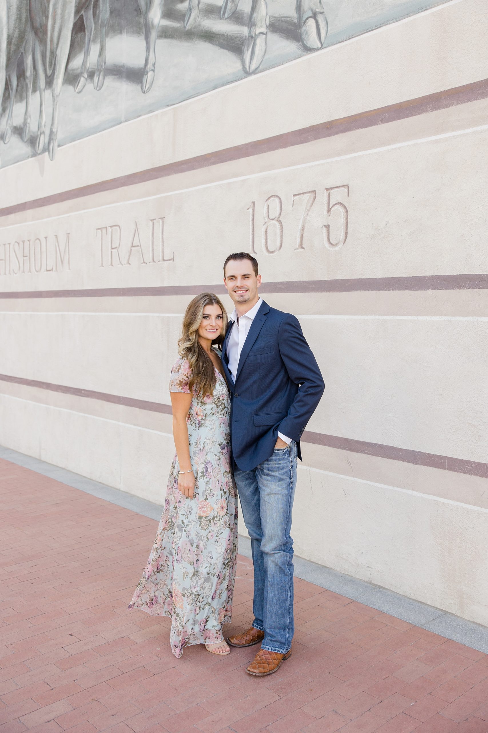 engaged couple hugs by tan wall at Chisholm Trail mural