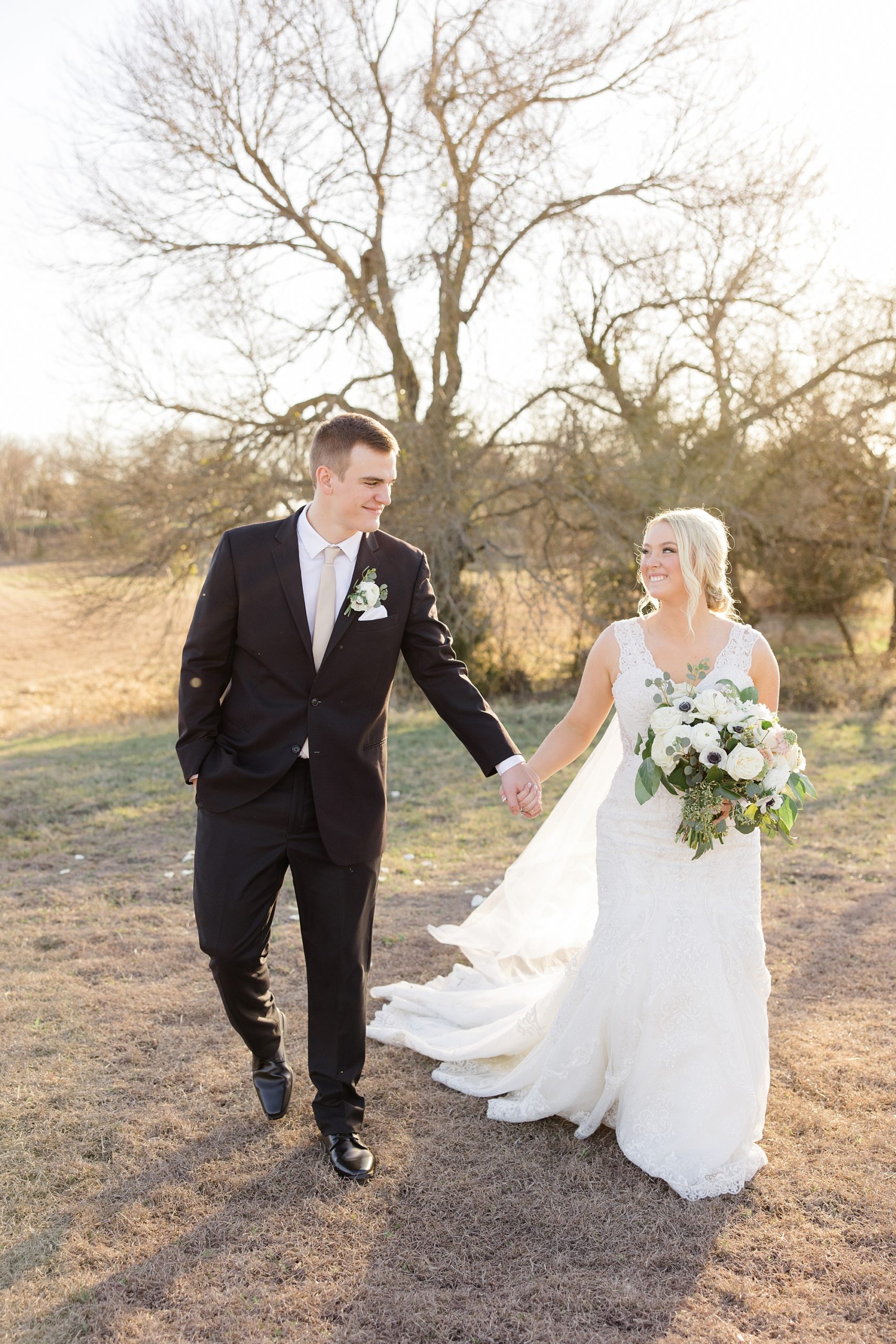 bride and groom hold hands walking through field at sunset 