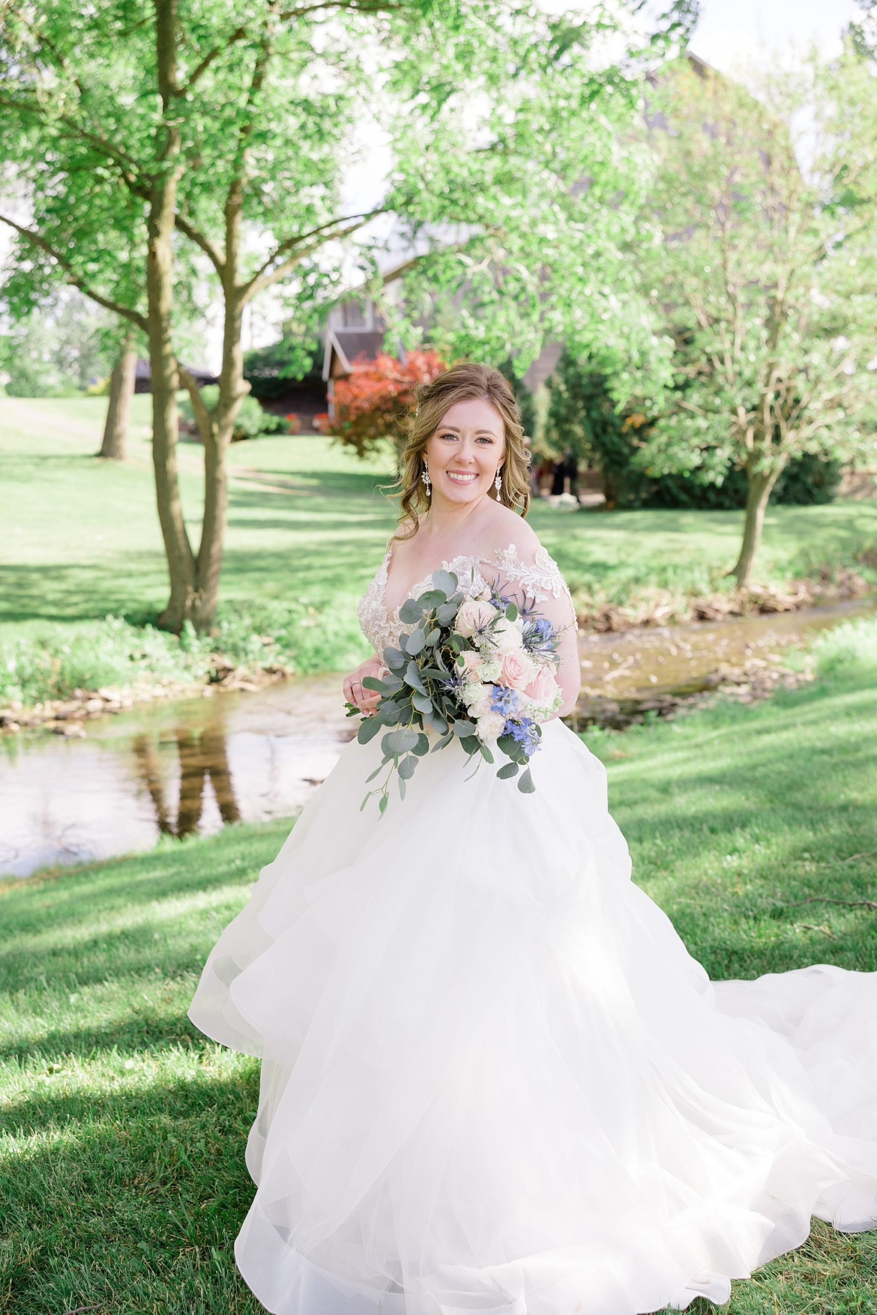 bride stands by creek at the Barn at Stoney Creek in wedding gown with lace sleeves