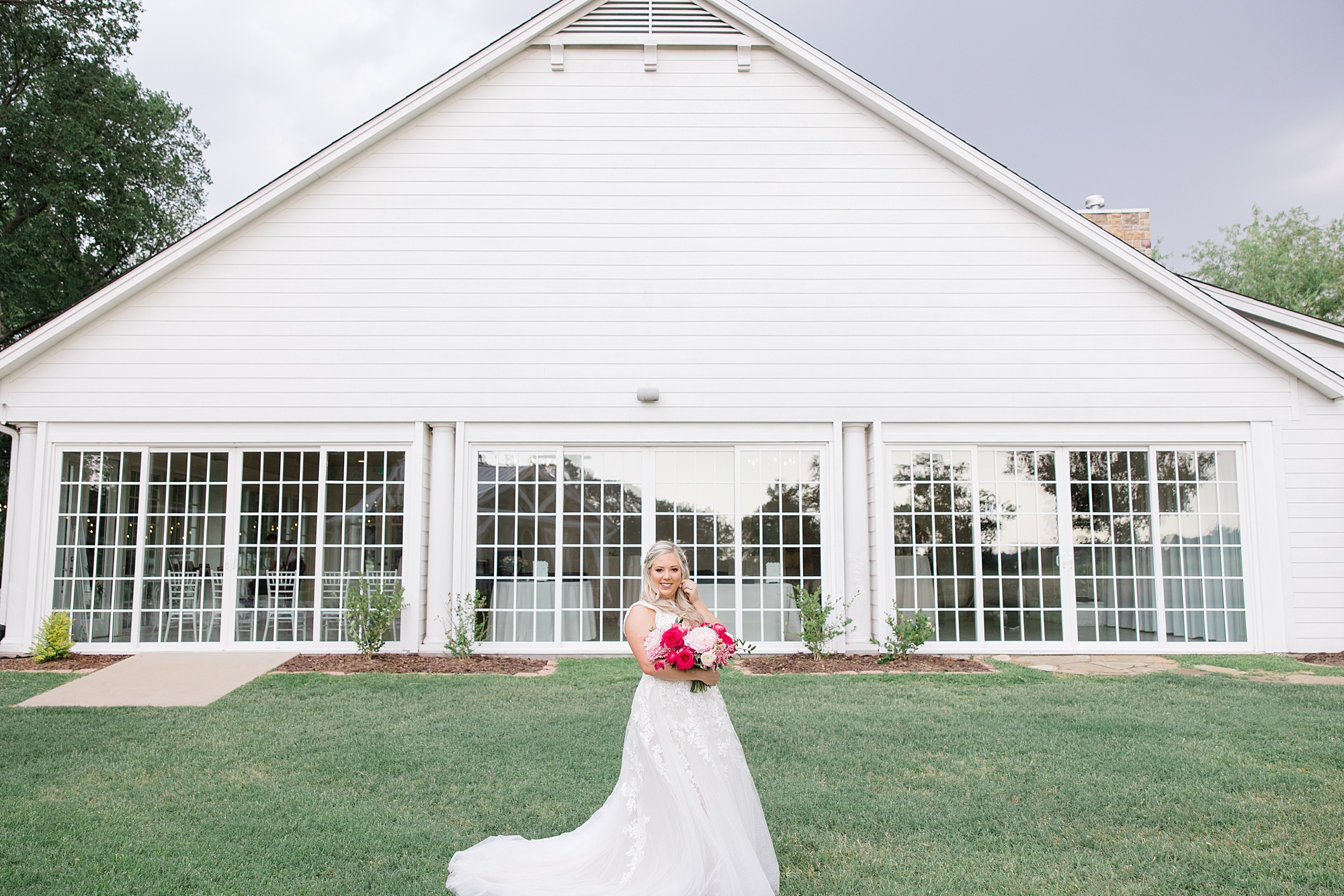 bride stands outside white building at Willow Creek with bright pink bouquet 