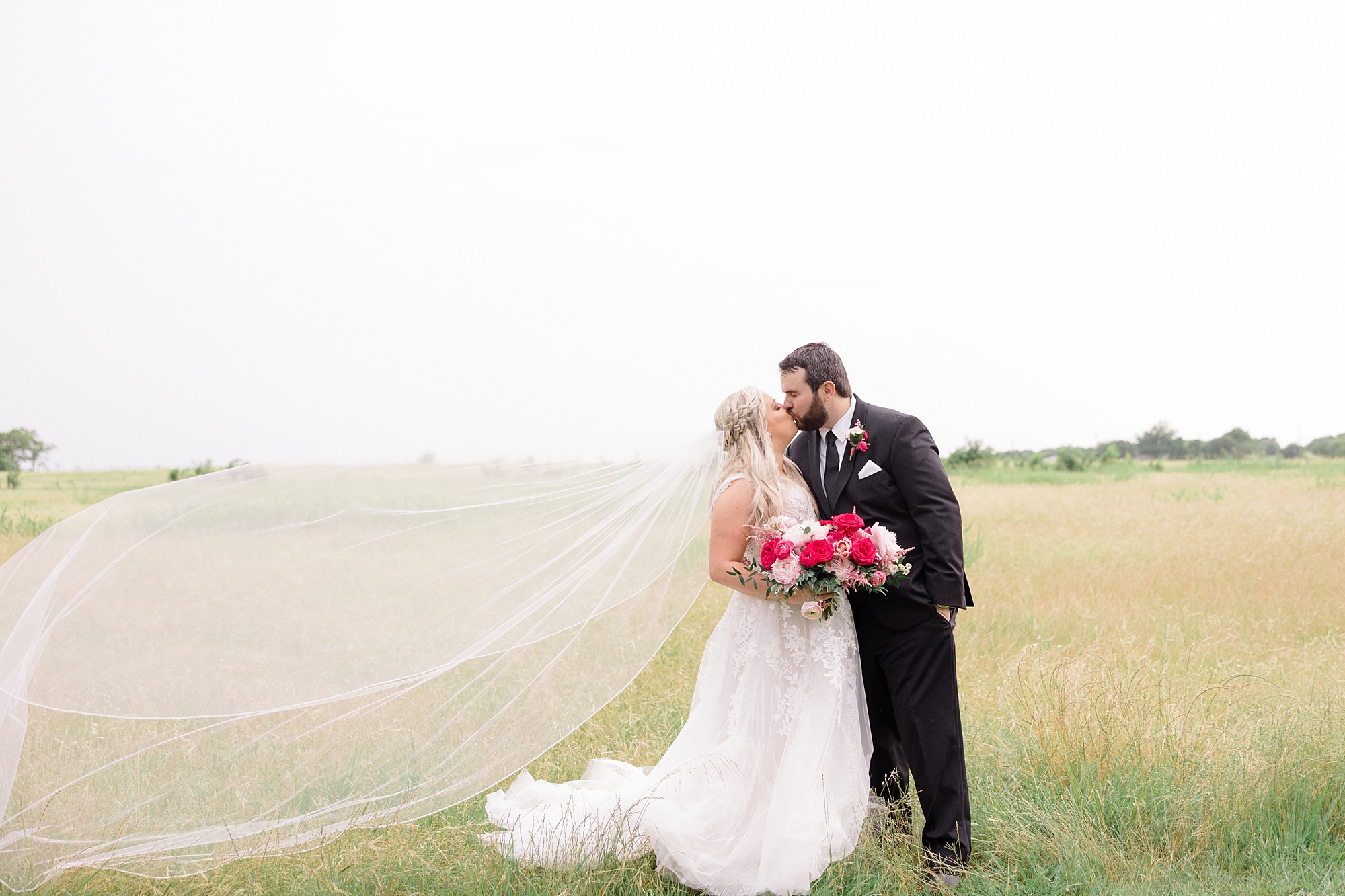 bride and groom kiss in field with bride's veil floating