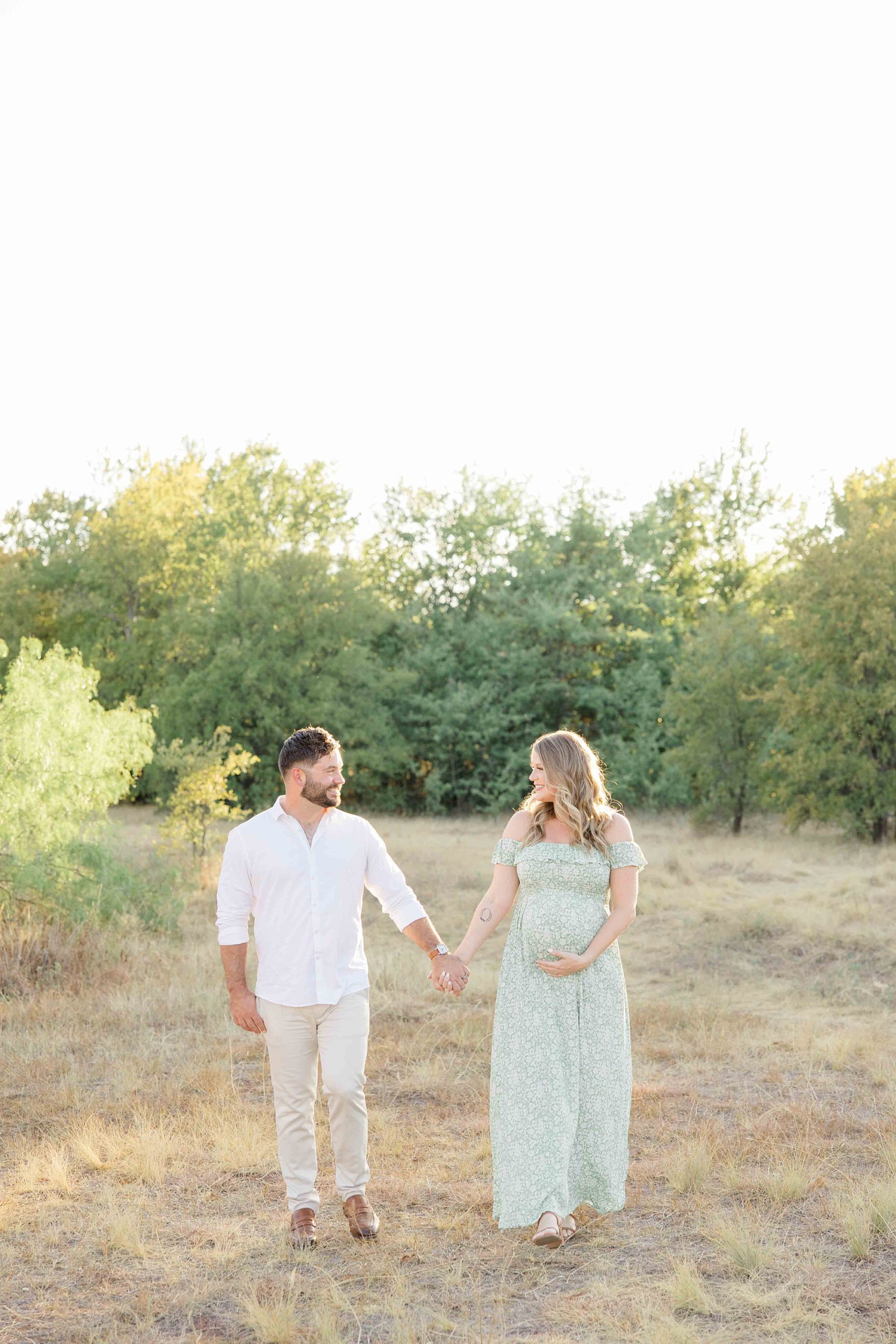 couple holds hands walking through field at sunset