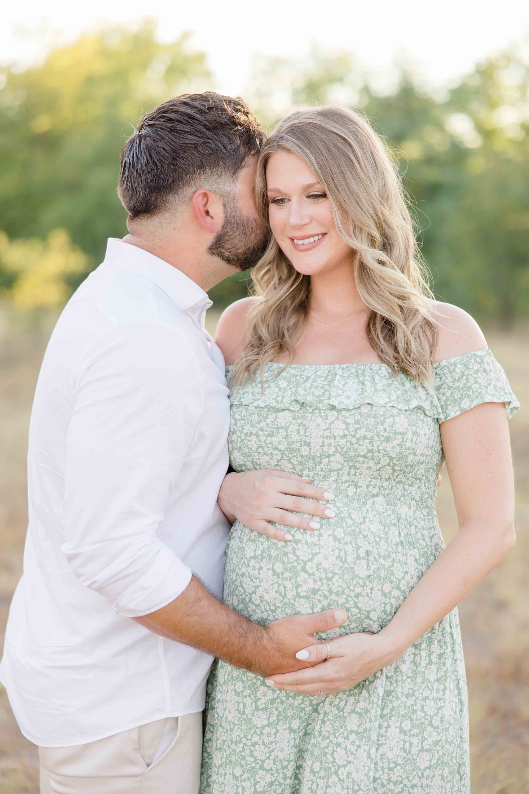 woman smiles while husband kisses her cheek while holding baby bump