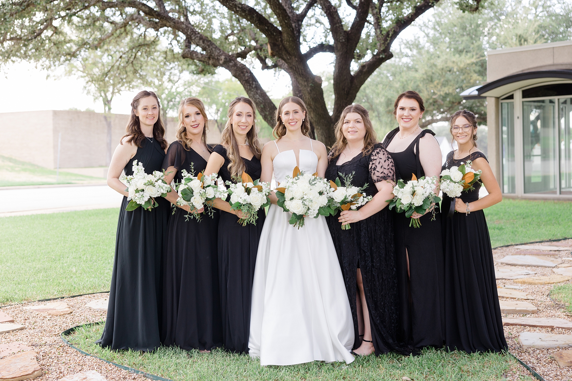 bride stands with bridesmaids in black gowns with ivory rose bouquets 