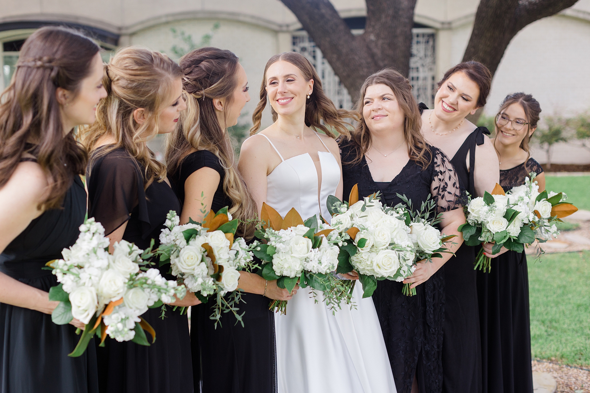 bride laughs with bridesmaids in mismatched black gowns with ivory rose bouquets 