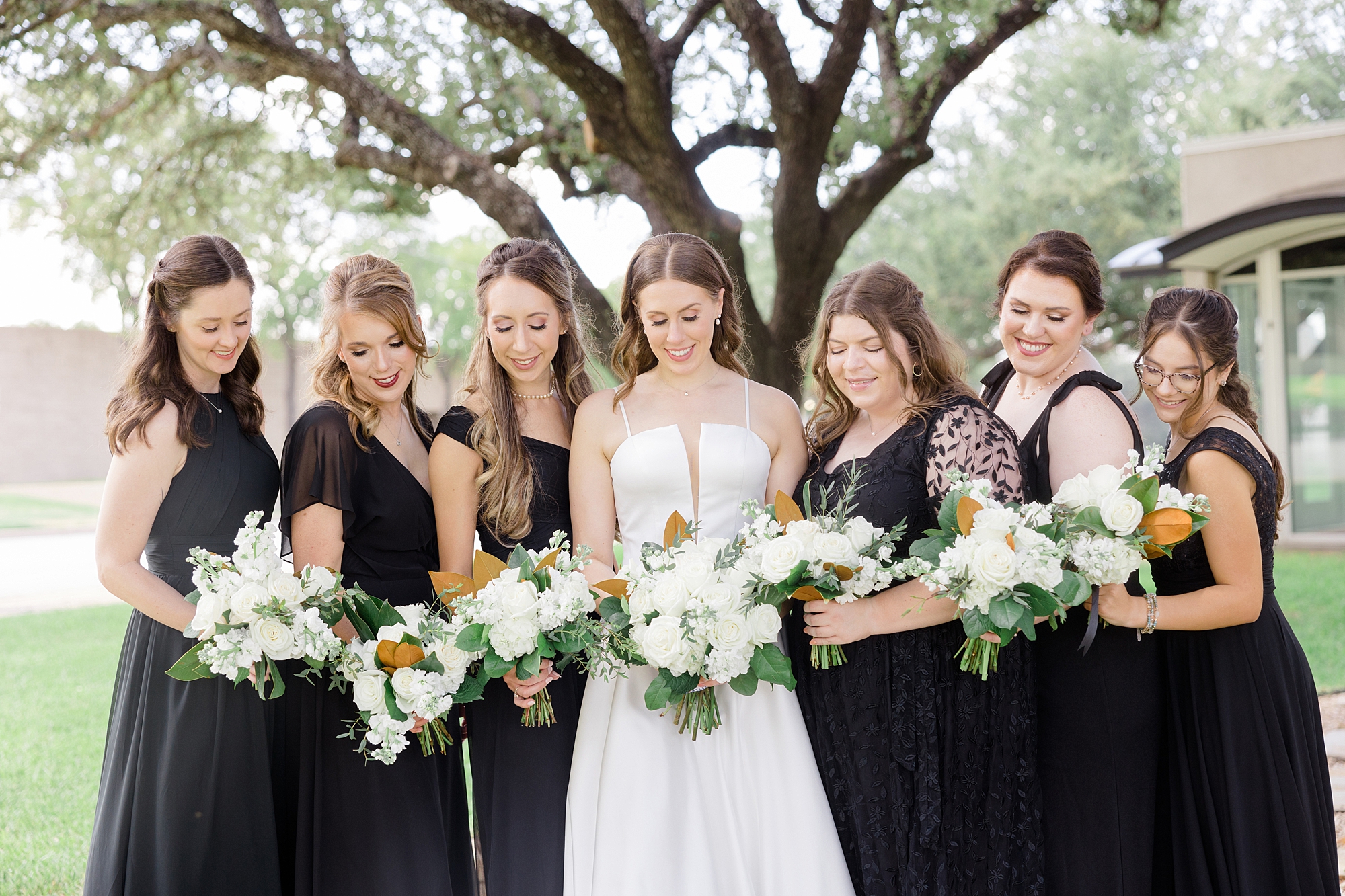 bride and bridesmaids in black gowns look down at ivory bouquets 