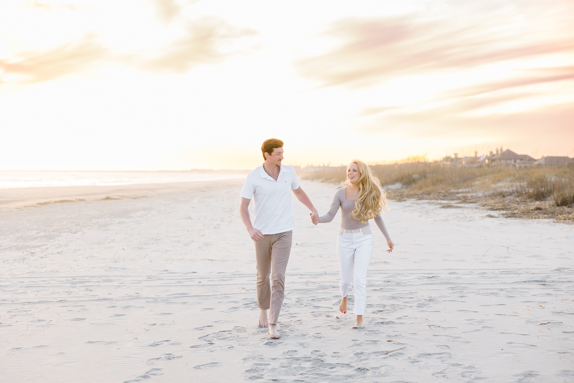 couple holds hands at sunset walking on sand during winter beach engagement session