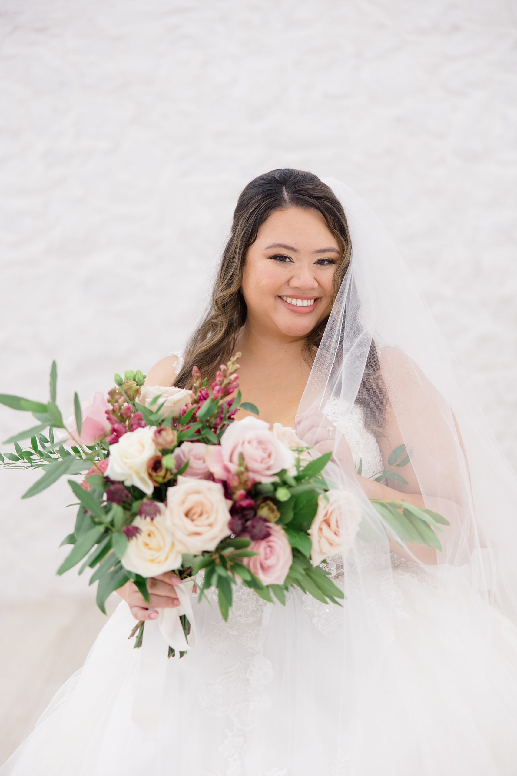 bride smiles with bouquet of pink flowers in from of her with veil around shoulders 