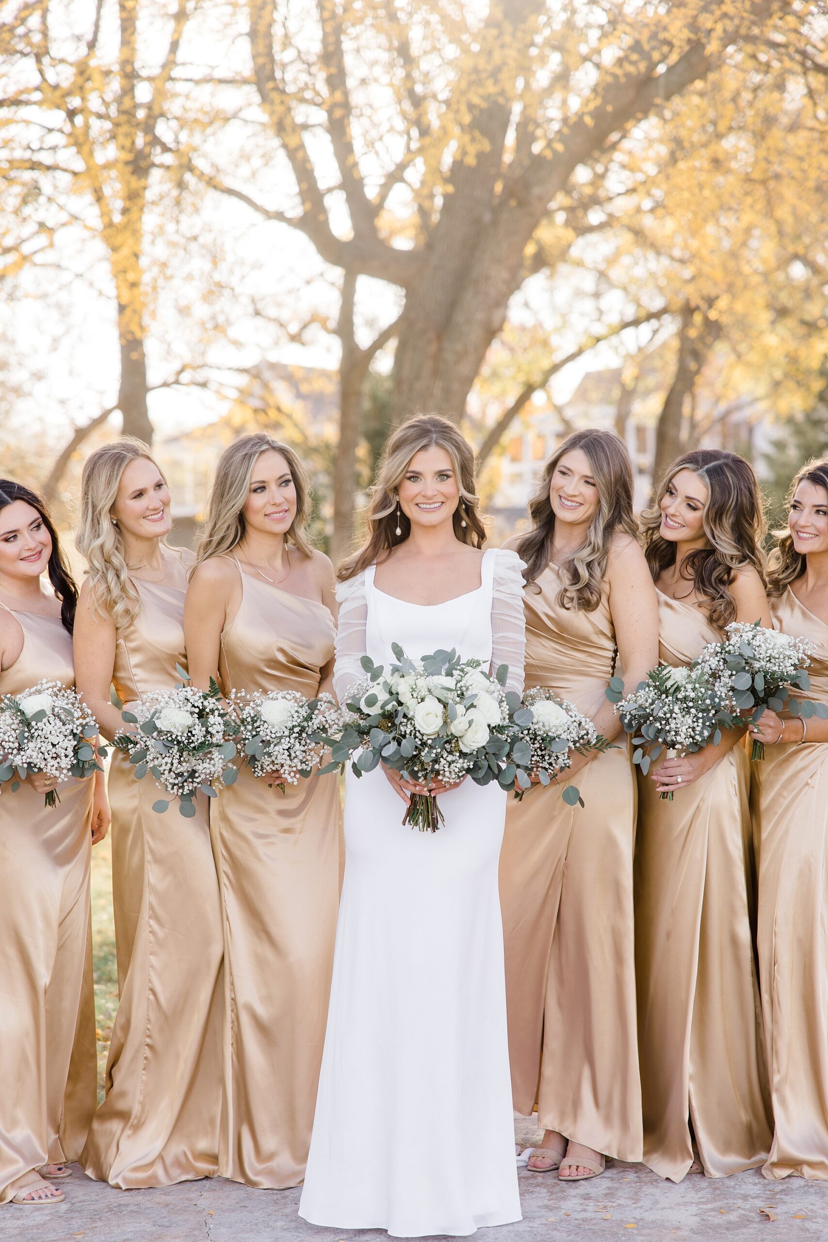 bride smiles standing between bridesmaids in gold gowns outside The Brooks at Weatherford