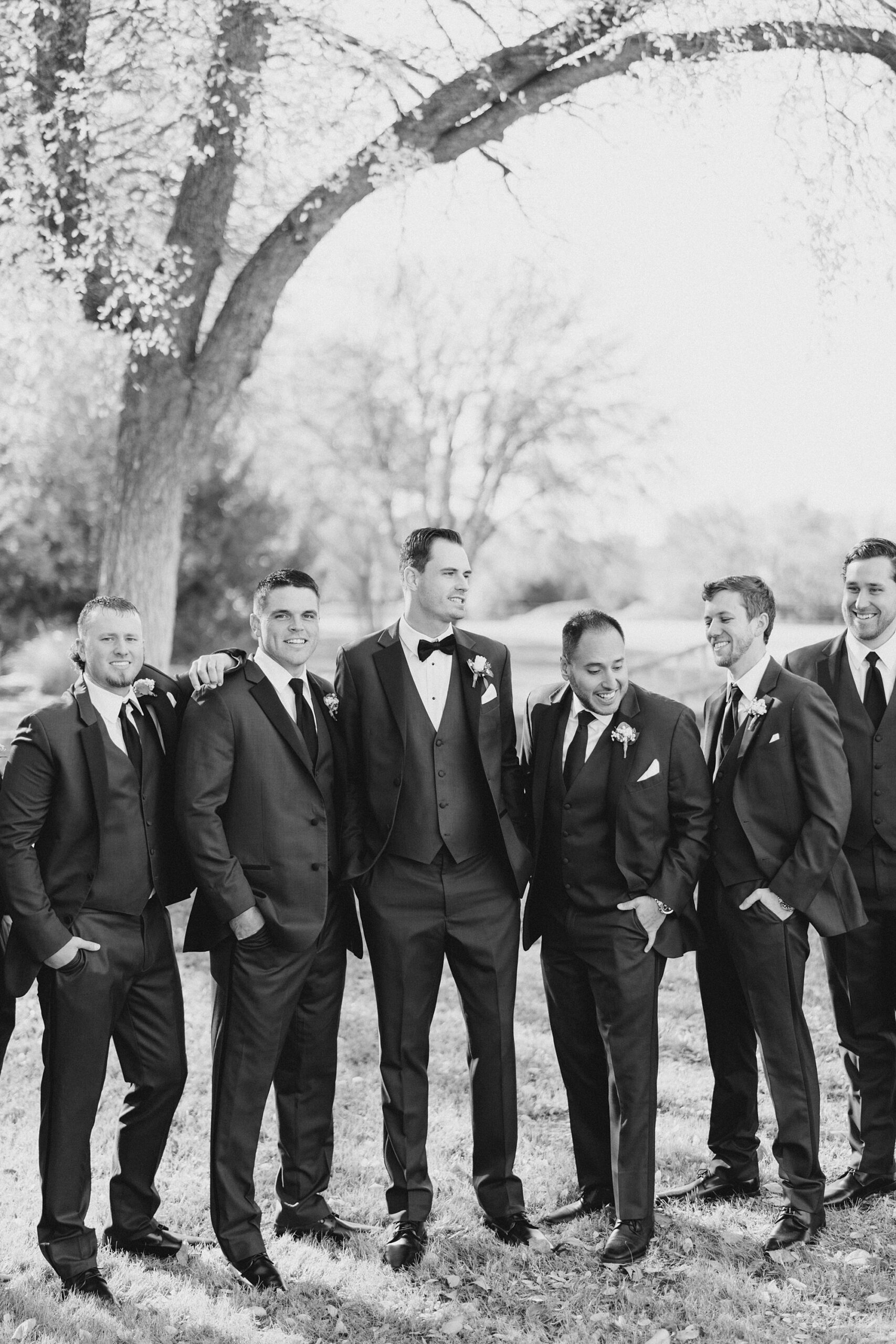 groom poses with groomsmen in suits under trees at The Brooks at Weatherford