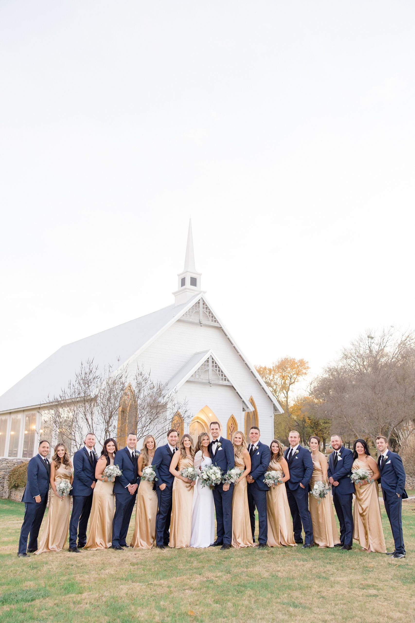 bride and groom pose with wedding party outside white chapel at The Brooks at Weatherford