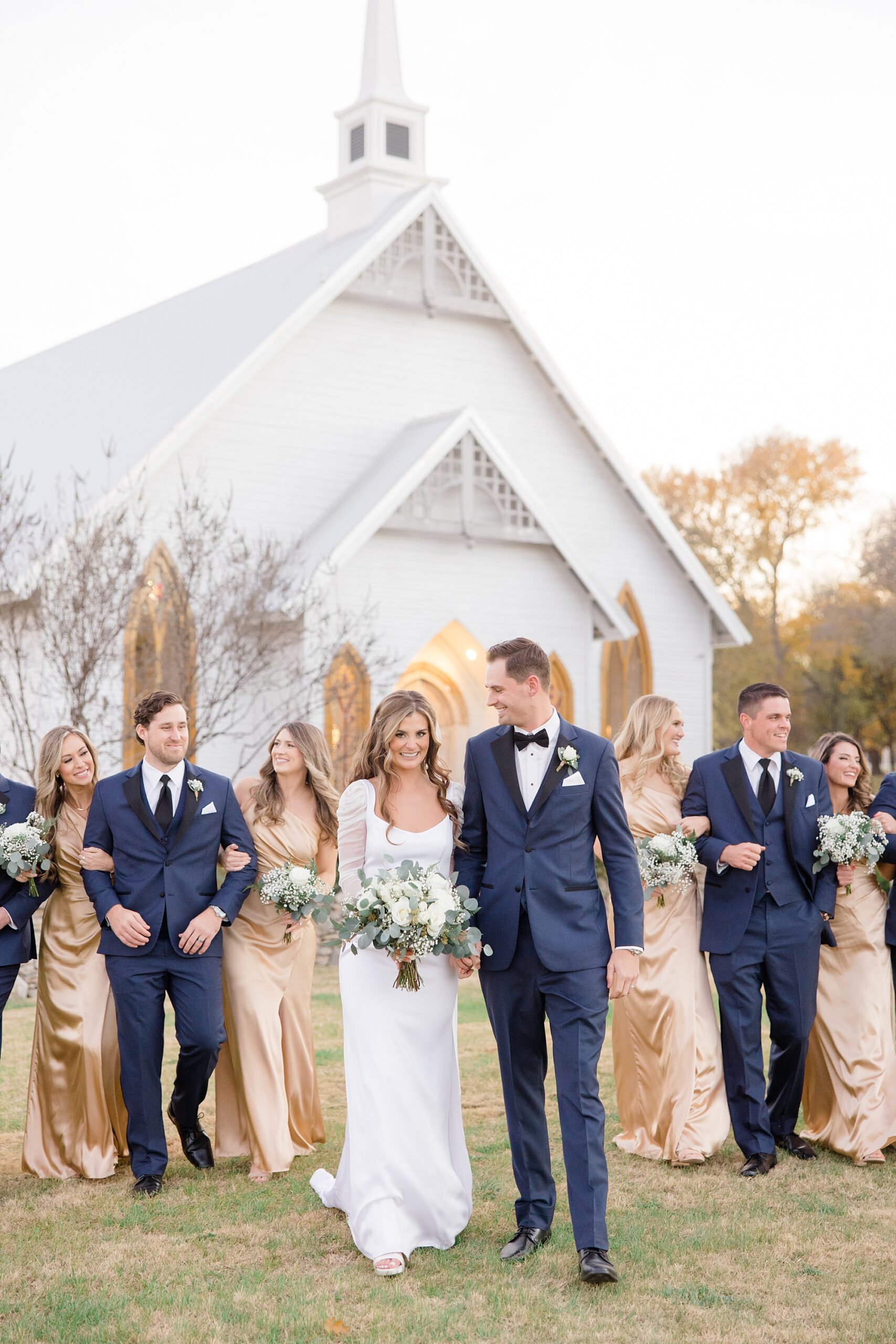 newlyweds walk in front of white chapel with wedding party behind them 