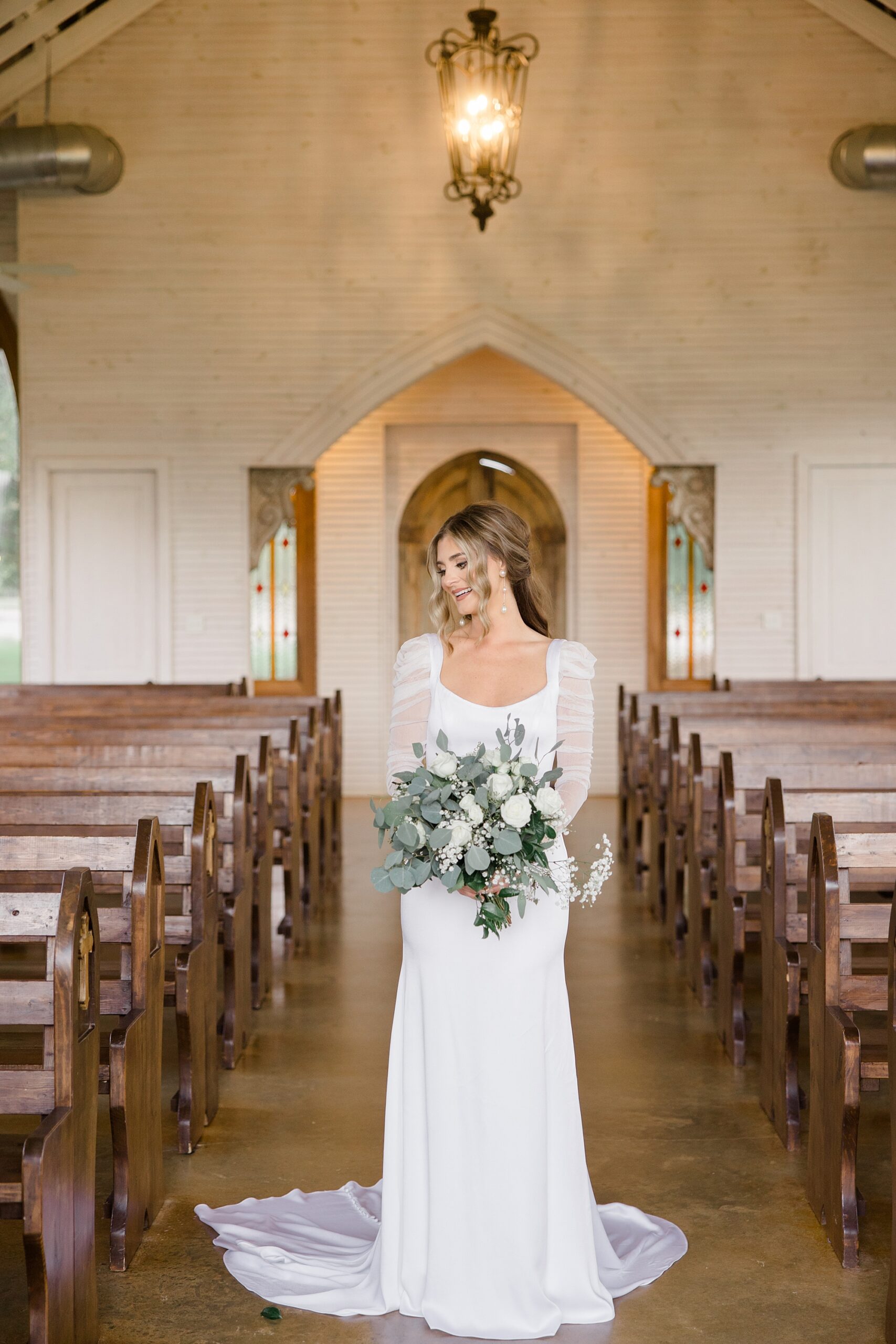bride holds bouquet of white flowers and eucalyptus leaves between pews in chapel at The Brooks at Weatherford
