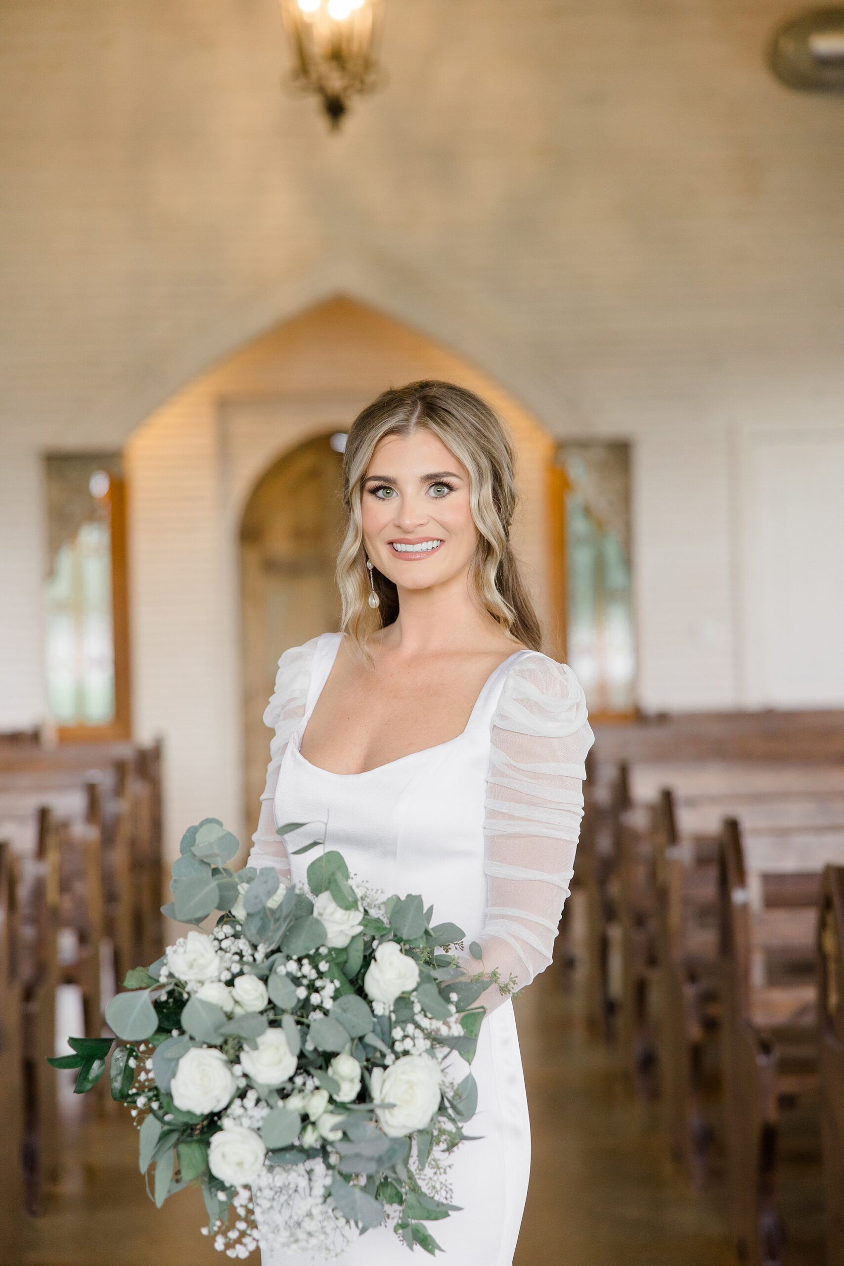 bride holds bouquet of white flowers and eucalyptus leaves between pews at The Brooks at Weatherford