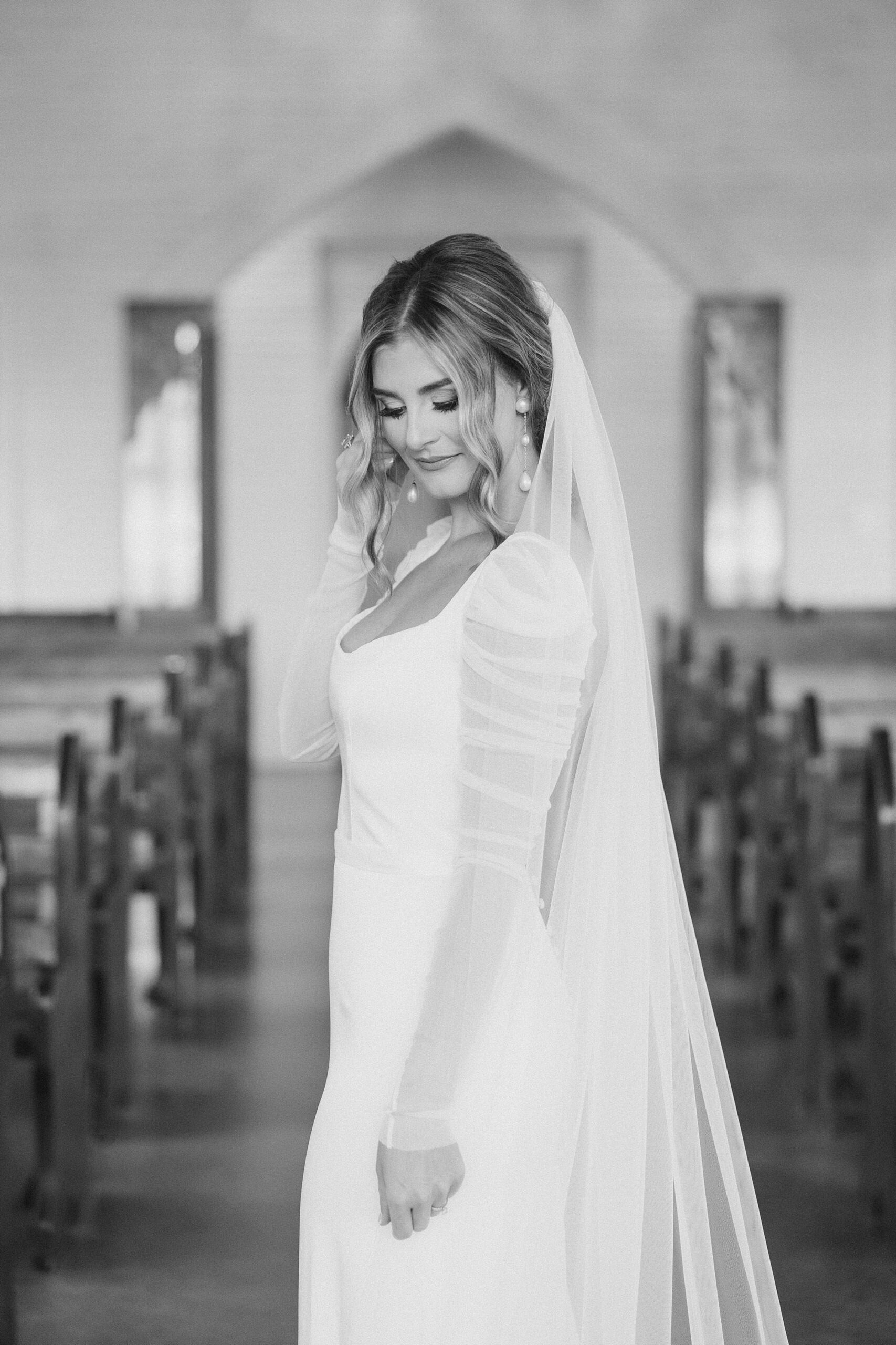 bride in long sleeve wedding gown with veil down back looks at floor between pews 