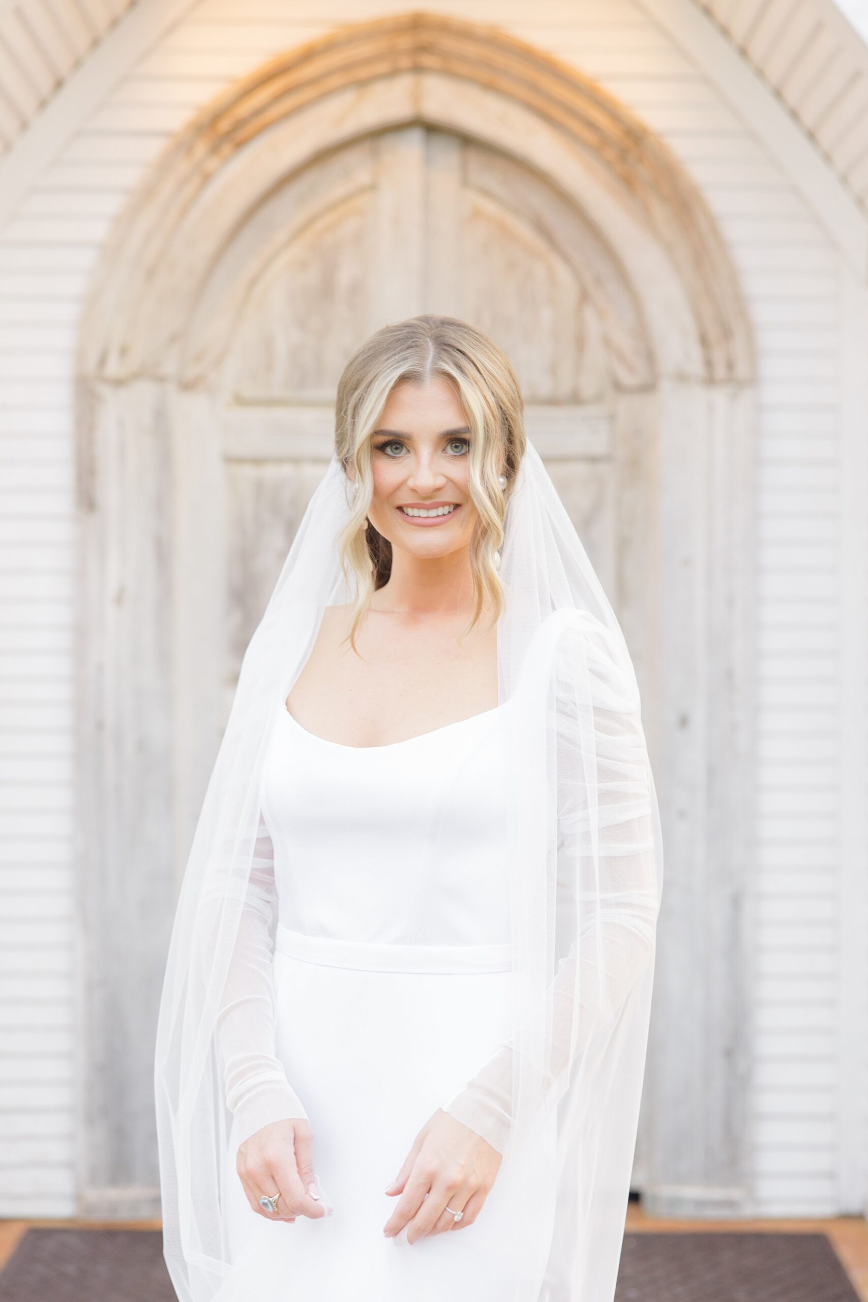 blonde woman in long sleeve wedding gown stands with veil around shoulders 