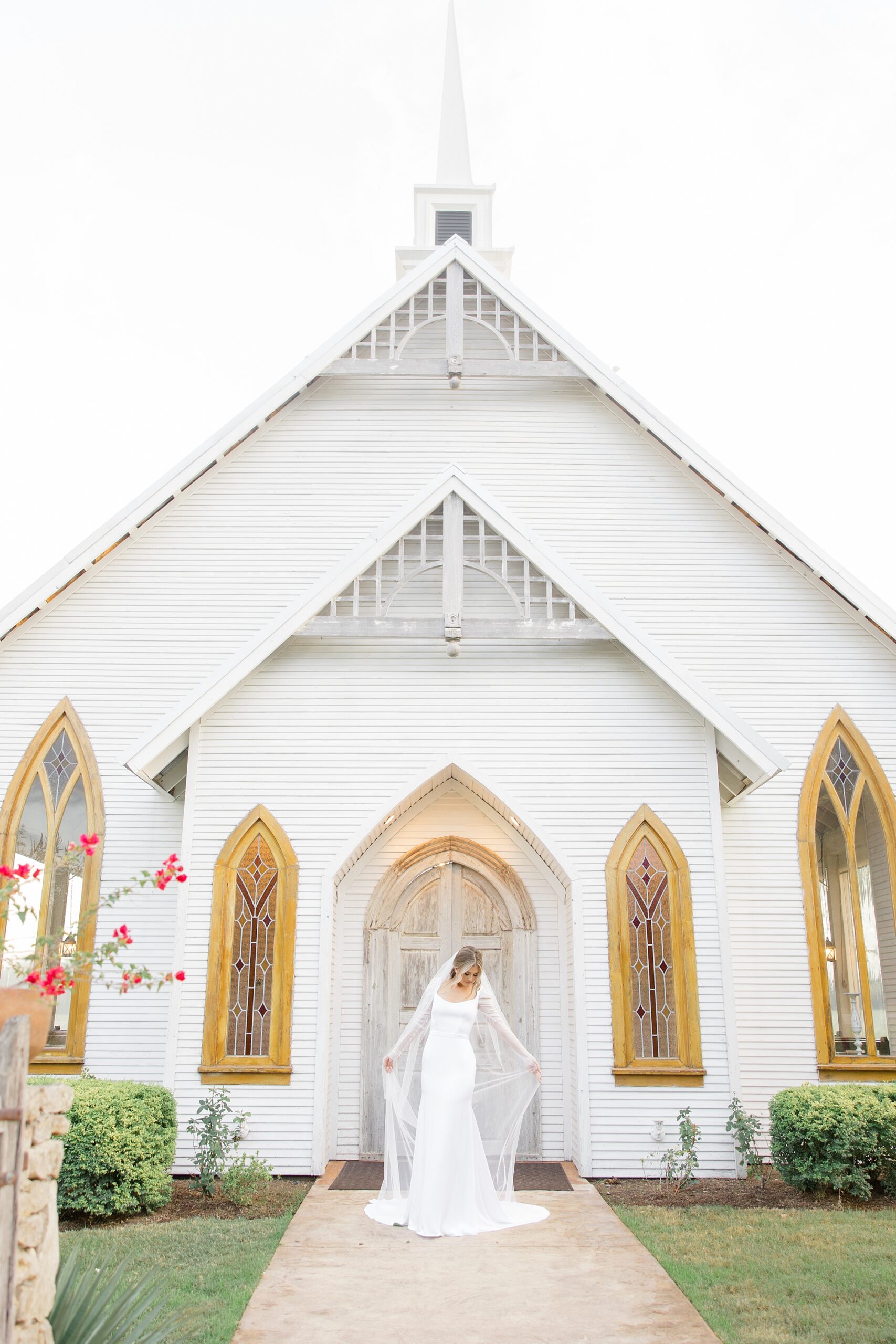 bride stands outside white chapel with veil being pulled around her 
