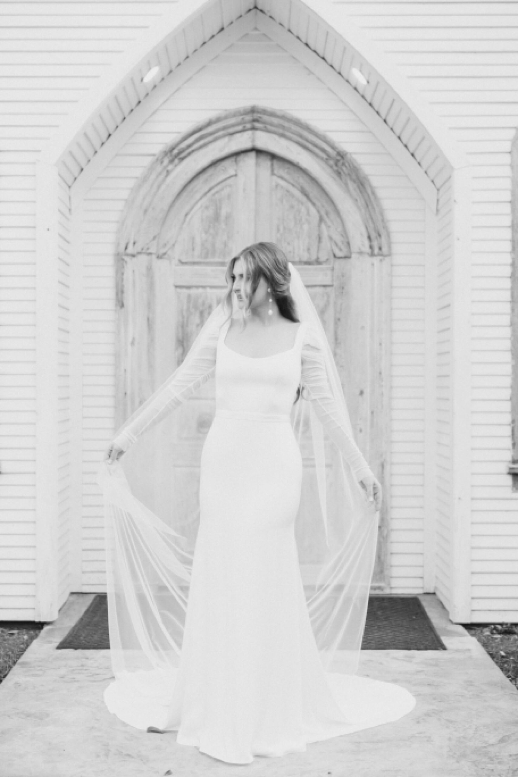 bride walks in front of wooden door holding veil out behind her 