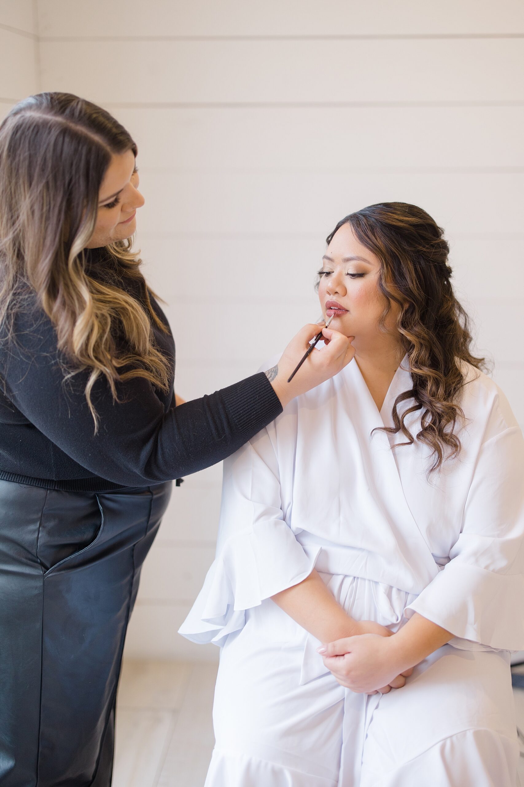 bride sits in white robe while makeup artist applies lipstick 