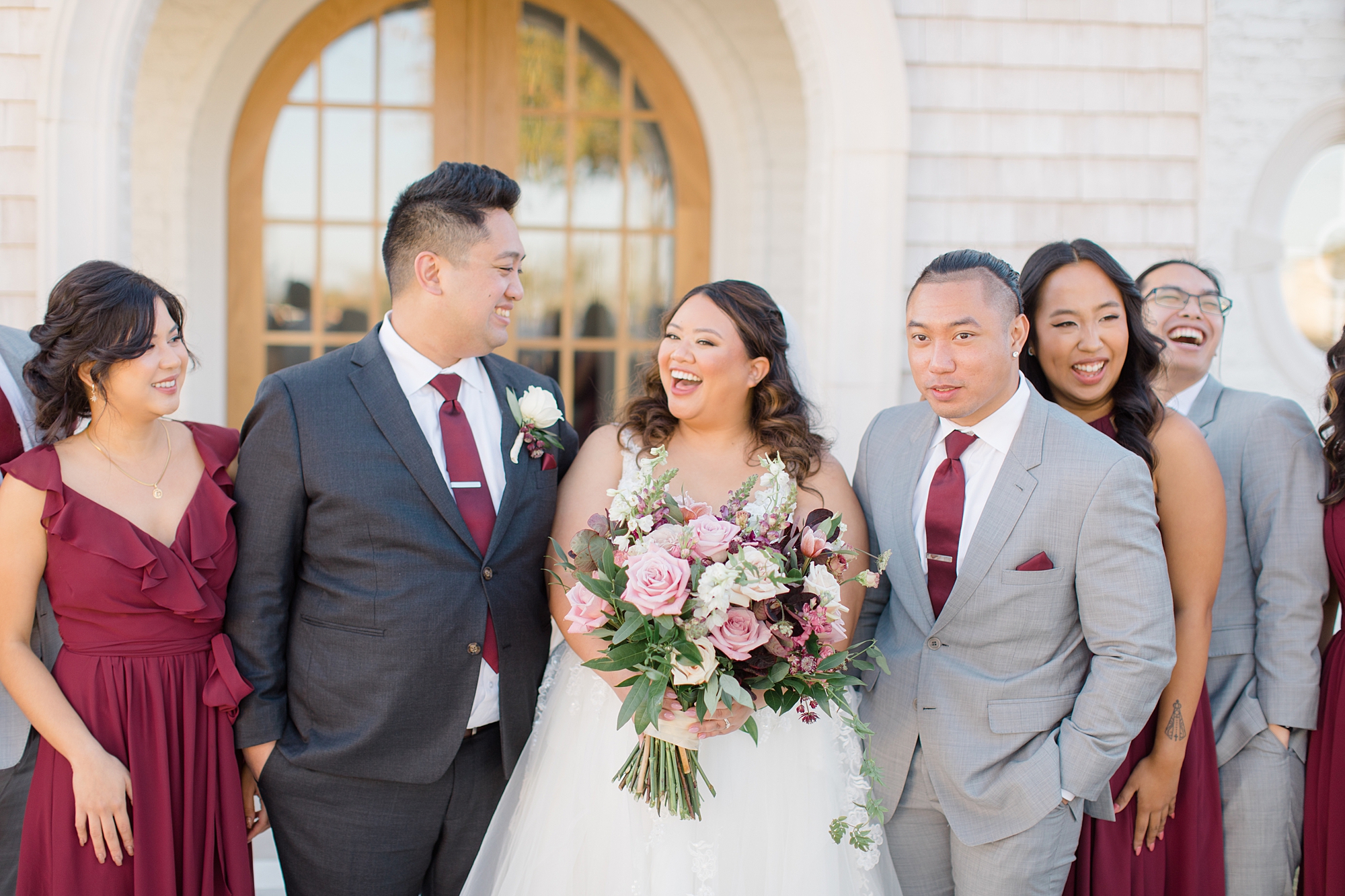 bride laughs with groom and wedding party in grey and burgundy 